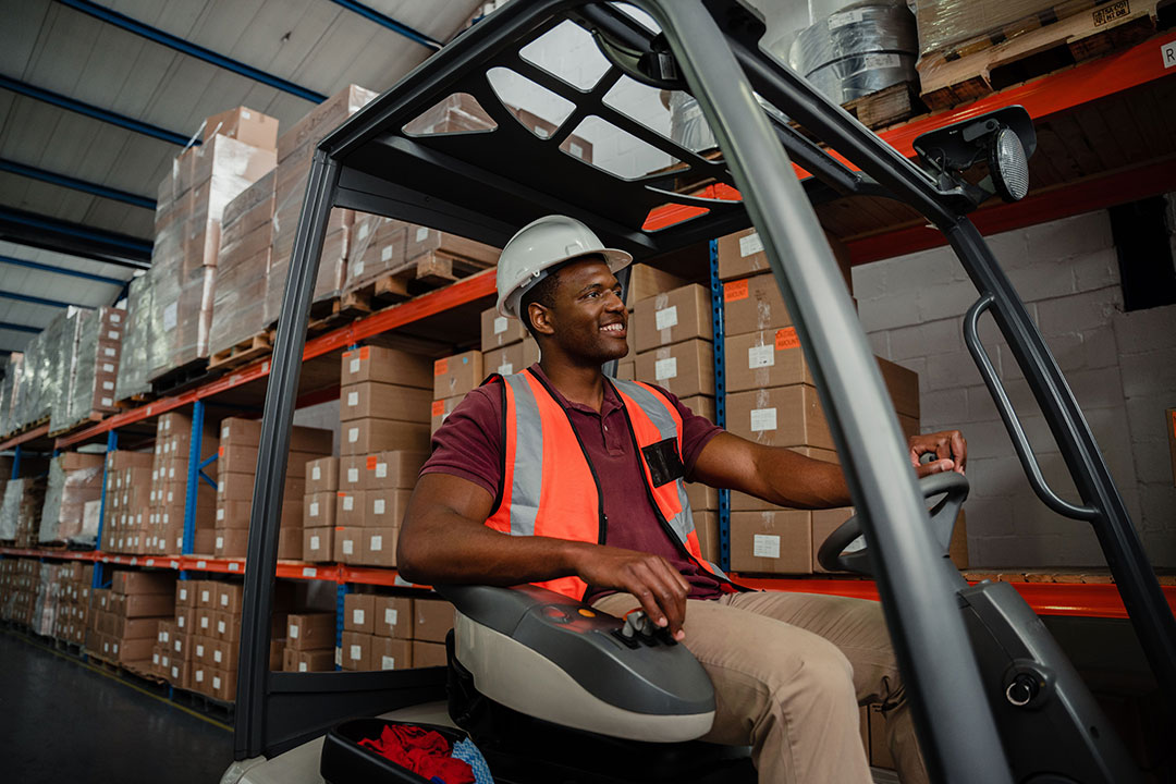 Mixed race worker driving forklift around warehouse while searching for parcels